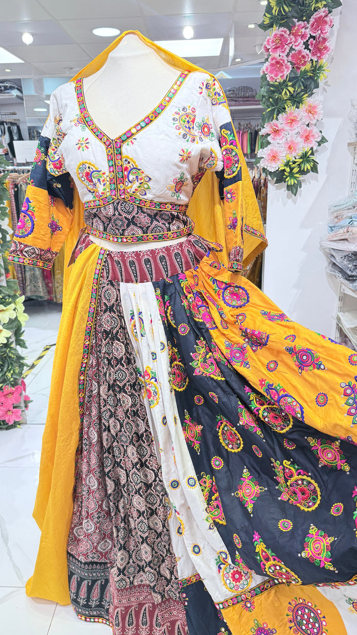 A multicolor traditional Indian outfit displayed on a mannequin in a retail store. The outfit features a white embroidered blouse with colorful patterns, a heavily patterned waistband, and a layered skirt in black, white, and bright yellow with intricate floral and paisley prints.